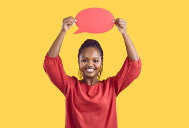 Portrait of happy Black woman standing isolated on yellow background and holding up empty red speech bubble. Studio shot of adult Afro American lady smiling and showing blank oval speech bubble card