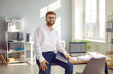 Portrait of young happy attractive bearded business man in white shirt and glasses sitting at the desk of his workplace with laptop with charts at the office, looking cheerful at camera and smiling.