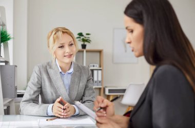 Two successful businesswomen sitting together in office, looking through paper documents, discussing future cooperation, have formal meeting, talking about new work project, reviewing deal