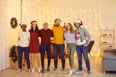 Pportrait of happy multiracial friends who gathered together at home to celebrate Christmas. People in santa hats standing in row hugging, smiling and looking at camera in room with christmas decor.
