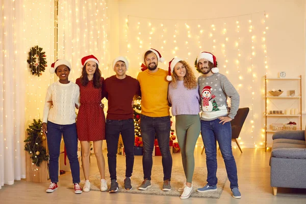 Pportrait of happy multiracial friends who gathered together at home to celebrate Christmas. People in santa hats standing in row hugging, smiling and looking at camera in room with christmas decor.