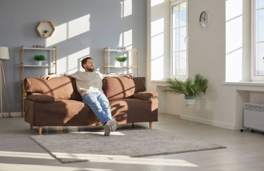 Happy house owner relaxing on comfortable sofa at home. Man enjoying leisure time and sitting on soft brown couch in cozy living room interior with white and light grey walls, shelves and green plants