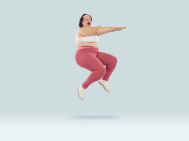 Full length photo of funny overjoyed young fat woman wearing sportswear jumping with excited expression, open mouth and closed eyes isolated on studio blue background. Workout, gym, fitness concept