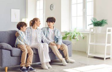 Happy family mother and two sons sitting together and hugging on sofa in living room. Young cheerful mom speaking with her children, laughing and smiling, enjoying home leisure time during weekends.