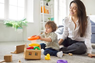 Young mother and son playing together, engaged in interesting game on floor, playing with pyramid, spending time at cozy home, having fun. Motherhood and child development concept.