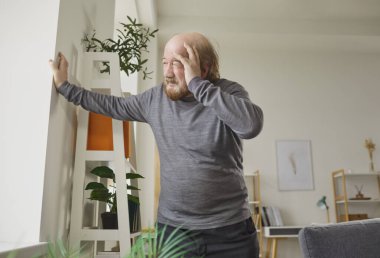 Dizzy senior man standing by window with hand on forehead and showing signs of pain or stress. Elderly male experiencing discomfort or anxiety, having vertigo, headache or blood pressure increase.