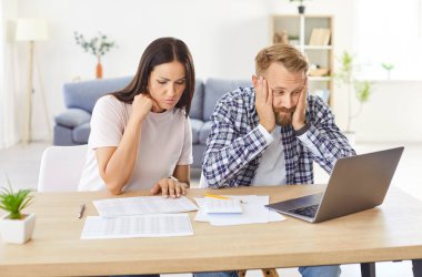 Sad focused young couple calculating bills in worry, discussing planning budget together, serious wife and husband looking at laptop screen, using online banking services and calculator, poor finances
