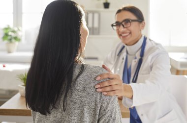 Portrait of smiling friendly doctor support female patient putting hand on her shoulder. Physician wearing stethoscope giving consultation a woman sitting back during medical examination in clinic.