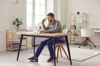 Focused bearded man working at home office with laptop computer. Serious young man successful freelancer wearing casual clothes sitting at desk looking at his laptop in front of him