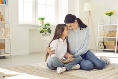 Mom spends time at home with daughter. Portrait of young mother and her teenage daughter who are sitting on floor in living room hugging and looking at each other with love. Mother and child concept.