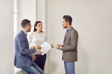 Group of office workers having a discussion. Team of three people talking about something while standing together by an office window. Work, business concepts