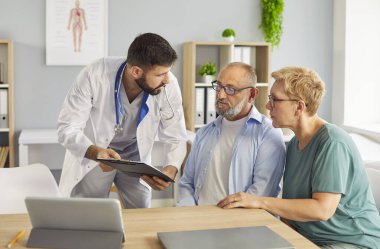Family doctor explains care plans to elderly patient and companion during medical consultation, discussing diagnosis. Male physician explains options to senior couple during exam in office.