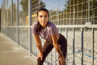 Kendine güveni tam genç, zayıf bir kadın açık hava koşusu sırasında soluklanmak için duruyor dizlerinin üstüne çöküp arka planda spor çitleri olan kameraya bakıyor.