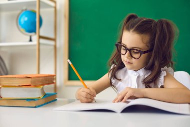 Back to school concept. A smart girl in glasses writes with a pencil in a notebook while sitting at a table in the classroom. Learning education lecture task equation for children in school.