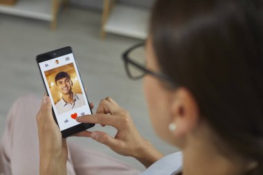 Close up of mobile phone with open dating application in hands of woman looking for relationship. Woman scrolling through profiles on dating site clicks red heart as sign of mans liking.
