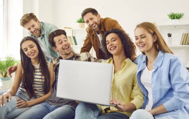 Friends using a modern laptop together. Group of happy young people sitting on the sofa at home, using a notebook computer, browsing internet, watching videos, talking and having fun