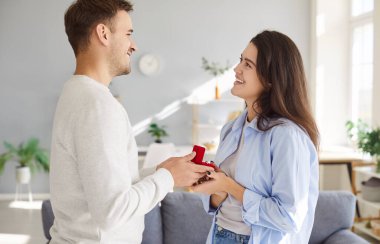 Young happy cheerful man giving red gift box to his girlfriend proposing marriage standing in living room at home. Male person giving surprise gift with engagement ring to pretty woman.