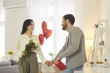 Cheerful young couple standing close, holding hands and smiling warmly at each other. Loving pair celebrating Valentine Day or special anniversary surrounded by flowers, balloons and gifts at home.