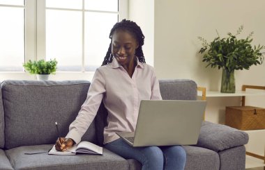 Portrait of a young happy smiling african american business woman working with laptop computer at home sitting on sofa and making notes in her notepad. Freelance and remote work concept.