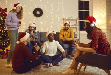Cheerful friends have fun together at home talking to each other and enjoying Christmas Eve. Happy people in santa hats sit with glasses of wine in cozy room with garlands and Christmas decorations.