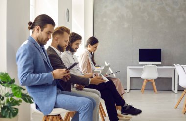 Diverse employees workers sit in row browse devices prepare for job interview in office. Multiracial people in queue use electronic gadgets ready for recruitment talk in company. Employment concept.