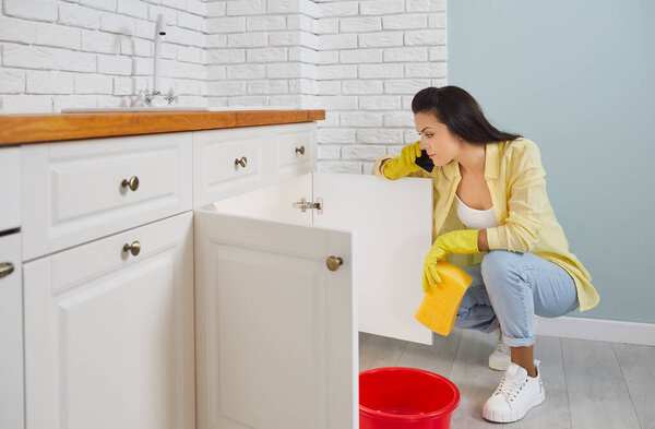 Frustrated woman on phone calls plumber because of leaking pipe in sink in kitchen. Young housewife in yellow rubber gloves sits near sink with bucket and washcloth and calls plumbing repair service.