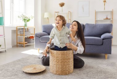 Young smiling mother having fun in the living room at home together with little cute child boy playing with basket. Smiling female parent spending time with son indoors. Happy family concept.