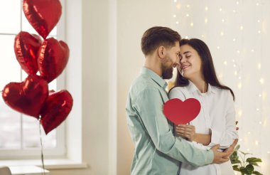 Smiling young man gave his heart to his attractive girlfriend on Valentines Day. Young couple in love in romantic atmosphere is sitting among heart-shaped balloons in the living room of the house.