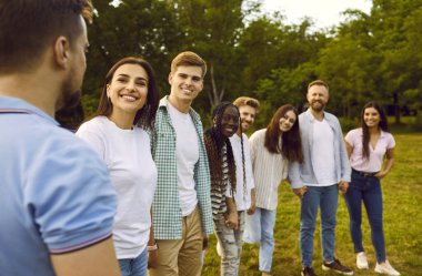 Happy young diverse friends meet in the park in summer. Several young people having fun in nature together. Group of cheerful, smiling, mixed race men and women standing in a row on a green grass lawn