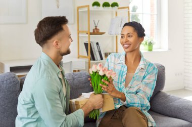 Young happy man holding present gift box and flowers in hand congratulating girlfriend on holiday. Smiling couple in love sitting at home celebrating anniversary. Valentines day concept.