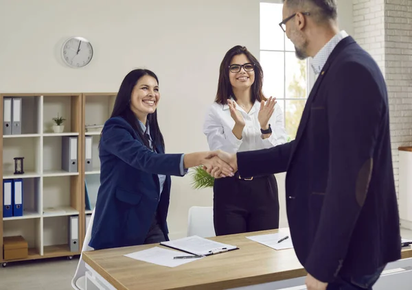 Handsome gray-haired businessman with glasses shakes hands with attractive brunette with long hair as sign of concluding good business deal. Positive atmosphere in office of three business partners.