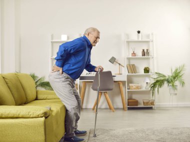 Senior man suffering from back pain at home. Elderly man with walking cane feeling pain while trying to get up from couch. Old age, medical and healthcare concept