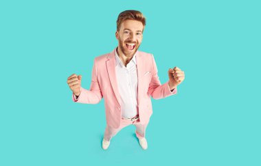 Happy guy celebrating his success and having fun. High angle shot of joyful young man in pink suit standing on blue color background, looking at camera, smiling and doing YES gesture. Fashion concept