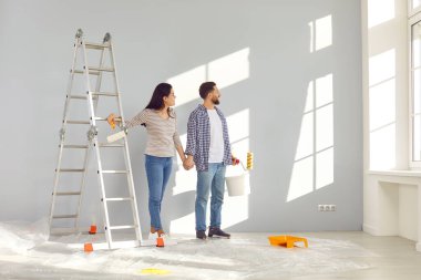 Smiling happy couple painting the wall of their new home holding paint rollers and looking at the window near ladder. Married man and woman doing repair renovation preparing to move into a new flat.