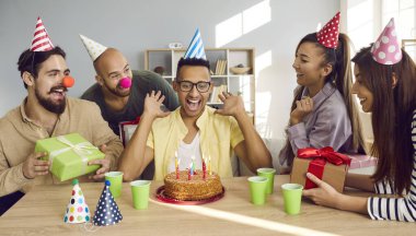 Overjoyed young African American man in celebration hat blow candles on birthday cake. Happy diverse multiracial people congratulate greet excited friend with anniversary. Party concept.