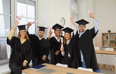 Congrats, grad. Group of happy international multiracial multicultural male and female university graduate students in graduation hats and robes standing in classroom, cheering and raising up diplomas