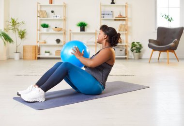 Happy African American woman doing abs exercises with a fitball at home gym on floor. Her training routine emphasizes fitness and sport, using gymnastic techniques to strengthen her core.