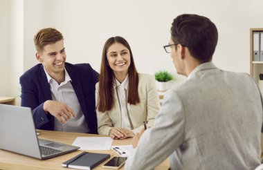 Happy young caucasian couple sitting at the desk and having consultation with man realtor or financial advisor in office. Business broker or insurance agent consulting man and woman in office.
