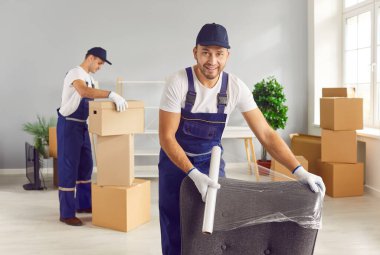 Movers male professional team in overalls loading, packing and unloading items with care, wrapping furniture in protective material, physically fit happy loader service men working 