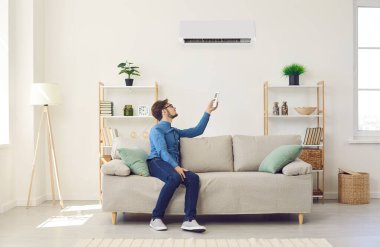 Young man turns on air conditioner with remote control while relaxing on couch at home. Man sits under air conditioning system and makes comfortable temperature for himself. Home comfort management.