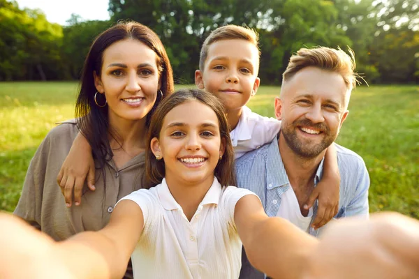 Happy family selfie in summer park. Parents and children having fun together. Pretty Caucasian european girl holds camera and takes photo of beautiful mother, handsome father and cute little brother