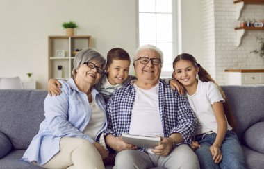 Portrait of a happy grandparents sitting on sofa with their grandchildren boy and girl at home with a family album and looking at camera. Kids enjoying leisure time with grandma and grandpa.