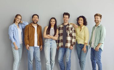 Team of people in modern casual wear standing by wall. Group of happy young men and women friends in comfortable shirts and jeans standing by grey wall, smiling and looking at camera all together