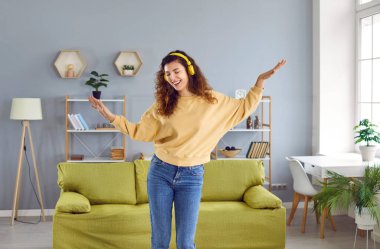 Happy woman listening to music at home. Beautiful young girl wearing a sandy sweatshirt, blue jeans, and yellow headphones enjoying good music, singing along, and dancing alone in the living room