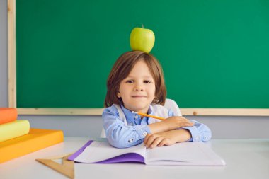 Serious boy sitting at desk with apple on his head in classroom. Adorable child going back to school. Elementary education concept