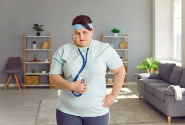 Obese, overweight young woman holding stethoscope on her big, fat belly and listening to bowel sounds caused by overeating and digestive system problems. Excessive weight, unhealthy lifestyle concept
