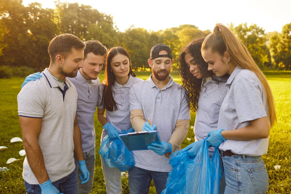 Group of young freinds, team adults gathering, looking at clipboard plan, holding blue bags, wearing gloves, reviewing list, collaborative, volunteering for cleanup, engaging in community action