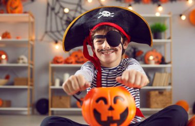 Indoor shot of happy child dressed up in kids pirate costume holding orange Halloween pumpkin basket. Portrait of smiling little boy with fake painted mustache wearing black hat with skull and bones