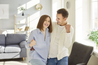 Home portrait of overjoyed happy young married couple in love posing, modern living room, yes gesture apartments, enjoy harmonic relationships, care and sincere feelings, celebrate success or victory 