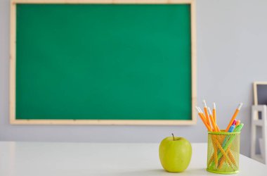 Sınıftaki öğretmenler masasında yeşil elma ve kırtasiye malzemesi tahtadaki metnin için boş yer. Meyve ve okul malzemeleri masada.
